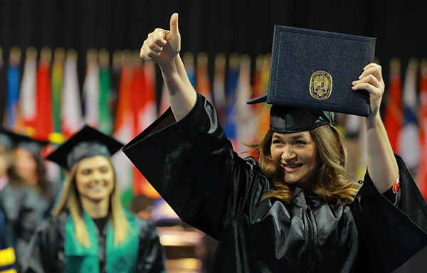 female student at graduation in cap and gown, holding up her diploma and giving the camera a thumbs up