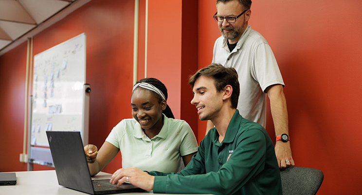 Two students sitting at a desk looking at a computer screen with a faculty member standing behind them also engaged